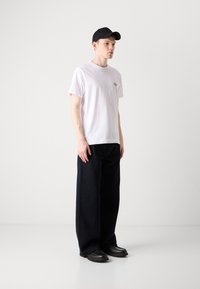 Hvid kortærmet T-shirt med et lille logo på brystet, kombineret med løstsiddende sorte bukser og sorte sko, båret sammen med en sort kasket.
