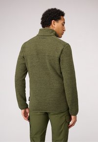 Giacca in pile di colore verde scuro con un motivo testurizzato, colletto alto e dettagli con logo sul retro. Abbinata a pantaloni cargo verdi.
