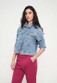 Τζιν μπουφάν σε ανοιχτό μπλε χρώμα με μακριά μανίκια, κλείσιμο με κουμπιά, δύο τσέπες στο στήθος και ακατέργαστο τελείωμα. Συνδυασμένο με ματζέντα παντελόνια ψηλότερης μέσης.