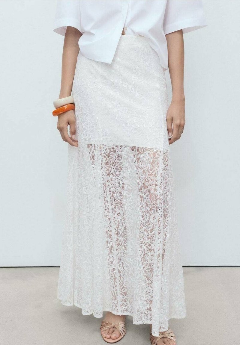 Femme portant une jupe longue en dentelle blanche avec une doublure opaque plus courte et une chemise blanche ample, accessoirisée avec des bracelets larges et des sandales beiges.