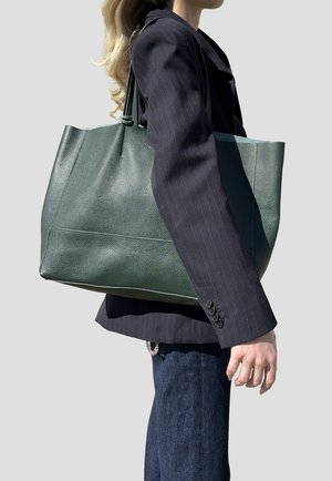 Donna con blazer a strisce sottili scuro e jeans, che porta una grande borsa tote in pelle verde scuro sulla spalla.