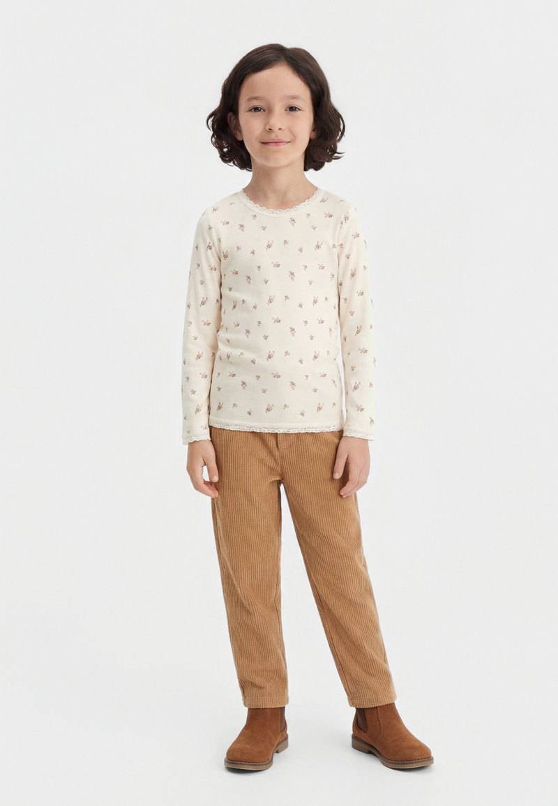 Enfant aux cheveux courts et bouclés, debout face à nous, portant une chemise crème à manches longues à fleurs, un pantalon en velours côtelé marron et des bottes marron.