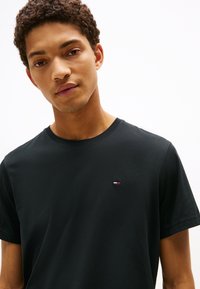 Juodas medvilninis marškinėlis su apvalia iškirpte, trumpomis rankovėmis ir mažu, išsiuvinėtu raudonos, baltos ir mėlynos spalvos logotipu kairėje krūtinės pusėje.