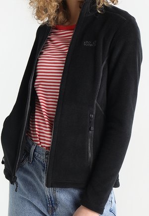 Persoon draagt een zwarte jack met ritssluiting over een rood-wit gestreept shirt en een blauwe spijkerbroek, staand tegen een effen achtergrond.