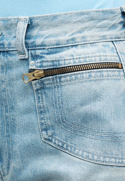 Φωτεινό γαλάζιο τζιν ύφασμα που δείχνει κοντινή εικόνα από την μπροστινή τσέπη με καφέ μεταλλικό φερμουάρ και θηλιά για ζώνη.