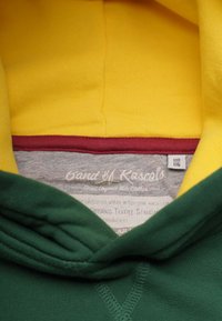 Huvtröja med grön kropp, gul insida på huvan och vinröd trim. Etiketten visar detaljer om ekologisk textilstandard. Mjuka tygtexturer.