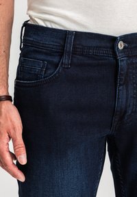 Mężczyzna ubrany w ciemnoniebieskie dżinsy i białą koszulę, z widoczną lewą ręką i brązową bransoletką po boku.