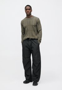 Πλεκτό πουλόβερ σε ελιά πράσινη απόχρωση με στρογγυλή λαιμόκοψη, σε συνδυασμό με μαύρο cargo παντελόνι και μαύρα αθλητικά παπούτσια, στέκεται μπροστά σε ένα απλό φόντο.