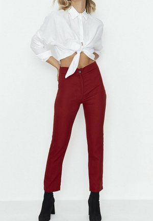 Kobieta w wysokim stanem burgundowych spodniach, białej, skróconej koszuli z guzikami, związanej w talii, oraz czarnych botkach do kostki, stojąca na białym tle.