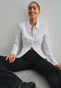 Bílá košile s dlouhým rukávem a špičatým límcem, přizpůsobená v pase, v kombinaci s černými kalhotami s vysokým pasem, na hladkém šedém povrchu.