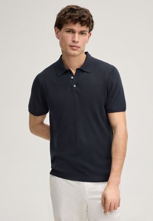 Giovane uomo con capelli ricci che indossa una polo blu navy a maniche corte e pantaloni bianchi, in piedi davanti a uno sfondo semplice.