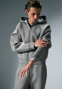 Harmaa vetoketjullinen huppari, joka on valmistettu pehmeästä kankaasta. Siinä on iso huppu, resorit hihansuissa ja rento istuvuus. Mallilla on sormissa sormuksia.