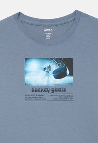 Szürke, rövid ujjú póló, amelyen egy hokis grafikája látható koronggal, alatta pedig különböző betűtípusokkal kísért szöveg található.