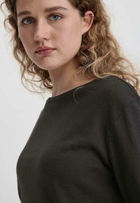 Dunkelgrüner, weicher Baumwollpullover mit Rundhalsausschnitt, mit lockerer Passform und leicht strukturierter Oberfläche, präsentiert an einer Person mit lockigem Haar vor neutralem Hintergrund.