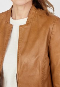 Bruine leren jas met een staande kraag, rits sluiting en een gladde textuur; gecombineerd met een lichtgekleurde gebreide trui eronder.