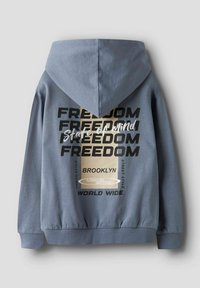 Hallituska värvi kapuutsiga, mille taga on suur graafiline trükk, mis sisaldab sõna "VABADUS" ja teisi teksti aktsente. Puuvillase segu pehme tekstuuriga.