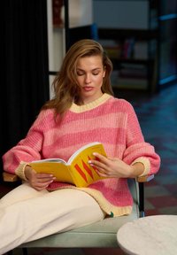 Przesadnie duży sweter z dzianiny w kolorze różowym z kremowymi wykończeniami, z poziomym wzorem w paski. Miękka tekstura, noszony z kremowymi spodniami, trzymając żółtą książkę.