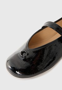 Black patent leather Mary Jane shoe with a rounded toe, decorative cutout detail, and adjustable strap. Smooth texture and minimal design.