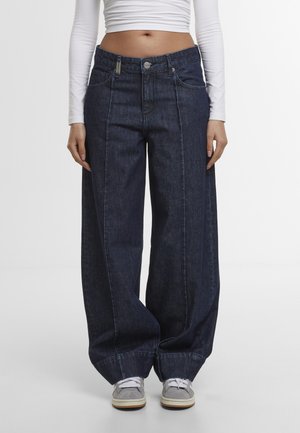 Nő sötét, széles szárú farmernadrágot, fehér, rövidített, hosszú ujjú felsőt és szürke sportcipőt visel, világos, egyszínű háttér előtt áll.