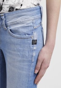 Vaqueros de mezclilla azul claro con un diseño desgastado, que cuentan con un corte elegante, cinco bolsillos y una etiqueta de marca visible en el bolsillo delantero.