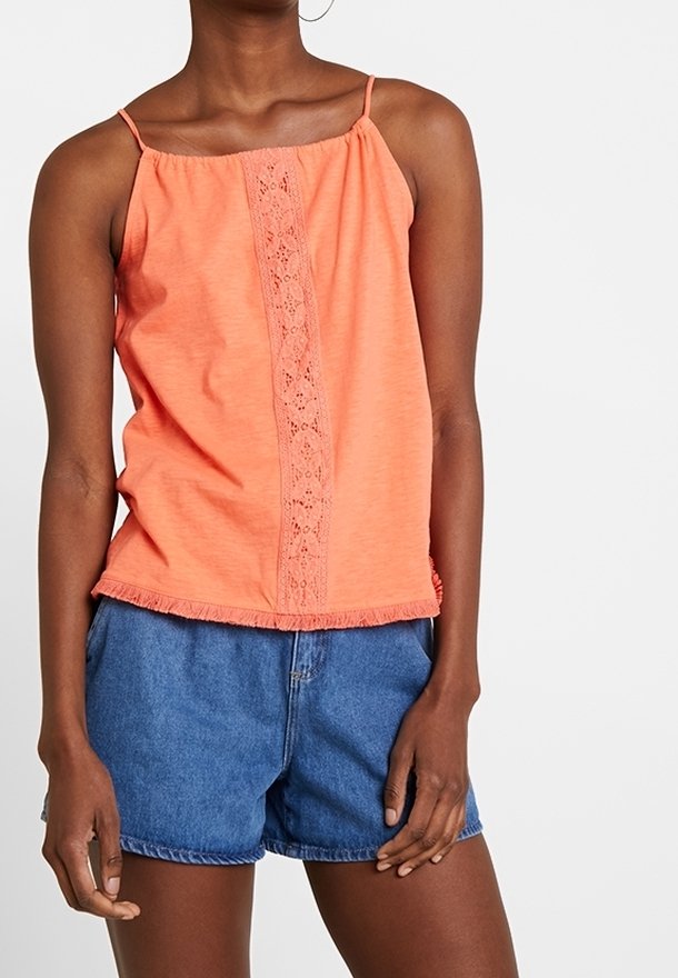 Femme portant un haut corail sans manches avec un détail en dentelle sur le devant et un short en jean bleu, debout devant un fond uni.