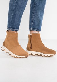 Brązowe zamszowe botki za kostkę z bocznym zamkiem i białą falistą podeszwą. Teksturowane wykończenie z miękką wewnętrzną wyściółką dla komfortu.