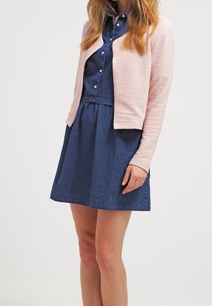 Vrouw die een marineblauwe jurk met knopen draagt met een taille die is geplooid en een lichtroze textuur vest met open voorkant.