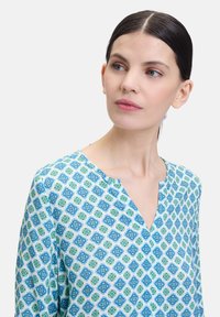 Patroonblouse met een blauwgroen geometrisch ontwerp op een witte achtergrond. V-hals, lange mouwen, lichte stoftextuur.