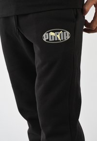 Pantalones deportivos negros confeccionados en una mezcla de algodón, con un logo ovalado amarillo y gris que representa a un gato saltando. Cintura elástica con cordón ajustable.