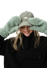 Jasnozielona kordowa czapka z logo serca; oversize fuzzy rękawice w tym samym odcieniu; czarna kurtka polarowa z wysokim kołnierzem.