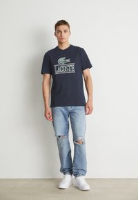 Camiseta azul marino con un logo de cocodrilo verde y el texto "LACOSTE" en blanco, combinada con jeans rotos de un azul claro y zapatillas blancas.