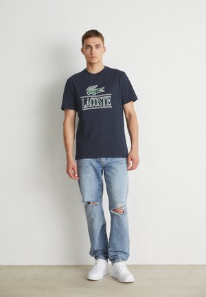 Dunkelblaues T-Shirt mit einem grünen Krokodil-Logo und weißem "LACOSTE"-Text, kombiniert mit hellblauen, ausgeblichenen Jeans und weißen Sneakers.