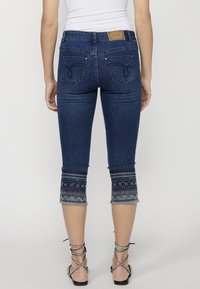 Jean capri bleu marine avec un ourlet effiloché, broderie décorative aux chevilles, et poches arrière avec des détails de couture subtils.