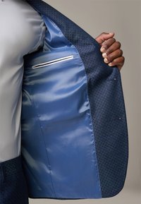 Interno di un giubbotto di abito blu navy su misura con una fodera in satin liscia, una tasca sul petto con bordino bianco e dettagli di tessuto texturizzato.