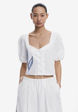 Top blanc court avec des manches bouffantes, un décolleté carré et des lacets sur le devant. Un motif de feuille brodé bleu ajoute des détails.