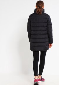 Mujer con cabello castaño en una coleta, vistiendo un abrigo acolchado negro, leggings negros y zapatillas rosas y negras, de espaldas sobre un fondo blanco.