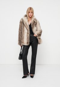 Chaqueta de piel sintética en tonos marrón claro, blusa negra y pantalones negros ajustados. Los accesorios incluyen un pequeño bolso negro y zapatos de punta.
