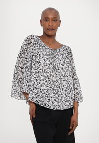 Svart og hvit blomstret bluse, gjennomsiktig stoff, løs passform, brede ermer, rynket halslinning med knytedetalj, lagd over mørke bukser.