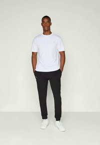 Witte katoenen t-shirt gecombineerd met zwarte sweatpants met zijzakken en een logo. Model draagt witte sneakers en staat op een lichte achtergrond.