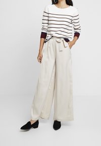 Sweter w paski w kolorze białym, bordowym i granatowym, zestawiony z luźnymi kremowymi spodniami satynowymi z wiązaniem w talii, noszony z czarnymi butami wsuwanymi.