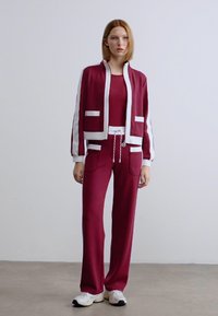 Μπορντό φόρμα με λευκές λεπτομέρειες, που περιλαμβάνει ένα μπουφάν με φερμουάρ και δύο μπροστινές τσέπες, καθώς και παντελόνι που ταιριάζει. Λείο, ελαστικό ύφασμα, άνετη εφαρμογή.
