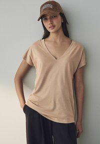 Beige V-ringad t-shirt, kort ärm, mjukt tyg, bärs med lösa svarta byxor. Brun keps med "AMOUR" i vita bokstäver.