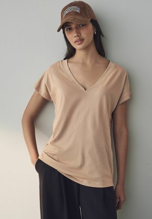 T-shirt beige à col en V, manches courtes, en tissu doux, porté avec un pantalon noir large. Casquette marron avec "AMOUR" en lettres blanches.