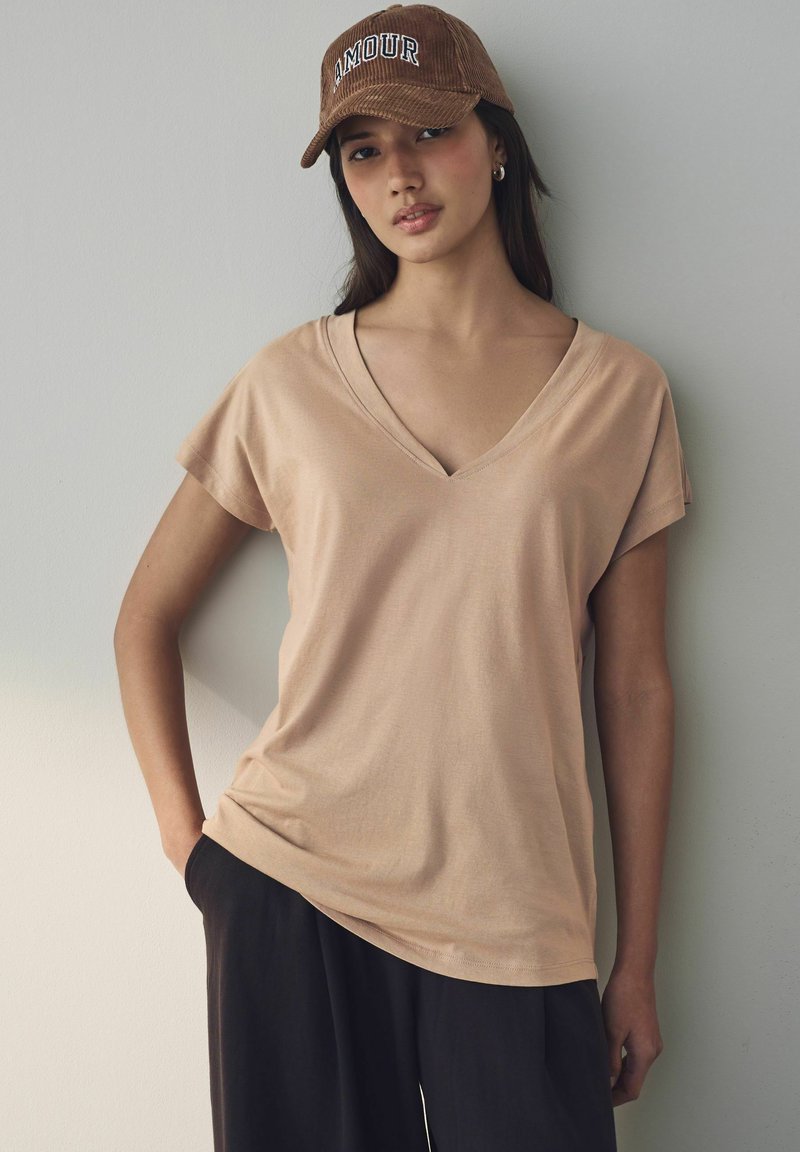 Beige V-ringad t-shirt, kort ärm, mjukt tyg, bärs med lösa svarta byxor. Brun keps med "AMOUR" i vita bokstäver.