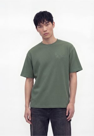 Jeune homme aux cheveux courts et foncés debout devant un fond uni, portant un T-shirt ample vert olive et un jean foncé, tourné vers l'avant.