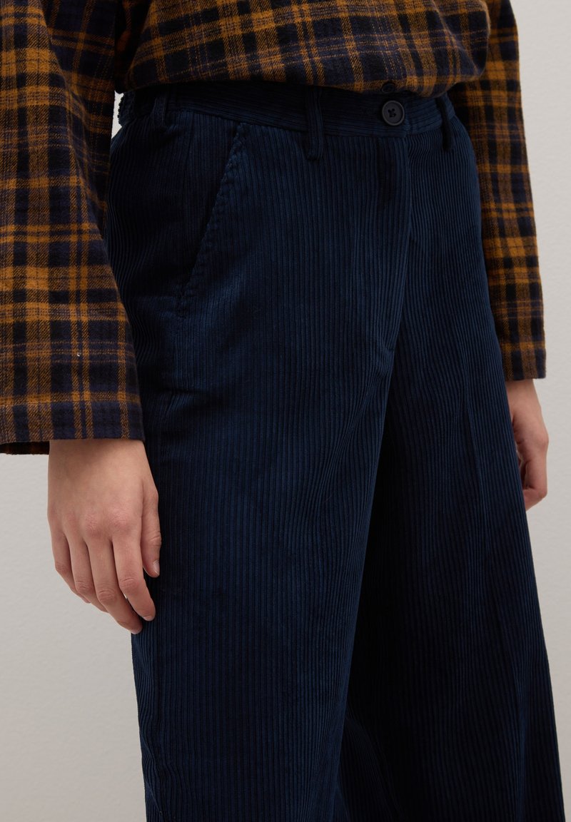 Marineblauwe corduroy broek met verticale ribbels, knoopsluiting en zijzakken, gecombineerd met een bruin-geel geruit shirt.