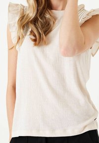 Donna che indossa un top senza maniche di colore crema con dettagli di spalle arricciate e pantaloni neri, in posa con un braccio sollevato.