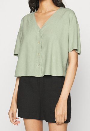 Personne portant un haut vert sauge à manches courtes boutonné avec col en V et un short noir taille haute sur un fond uni.