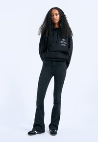 Black hoodie with large kangaroo pocket and graphic print, paired with black flared jeans and black shoes with white accents, on a plain background.