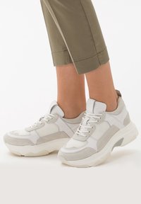 Para białych i jasnoszarych sneakersów z grubymi podeszwami, noszonych z krótkimi oliwkowymi spodniami na białym tle.